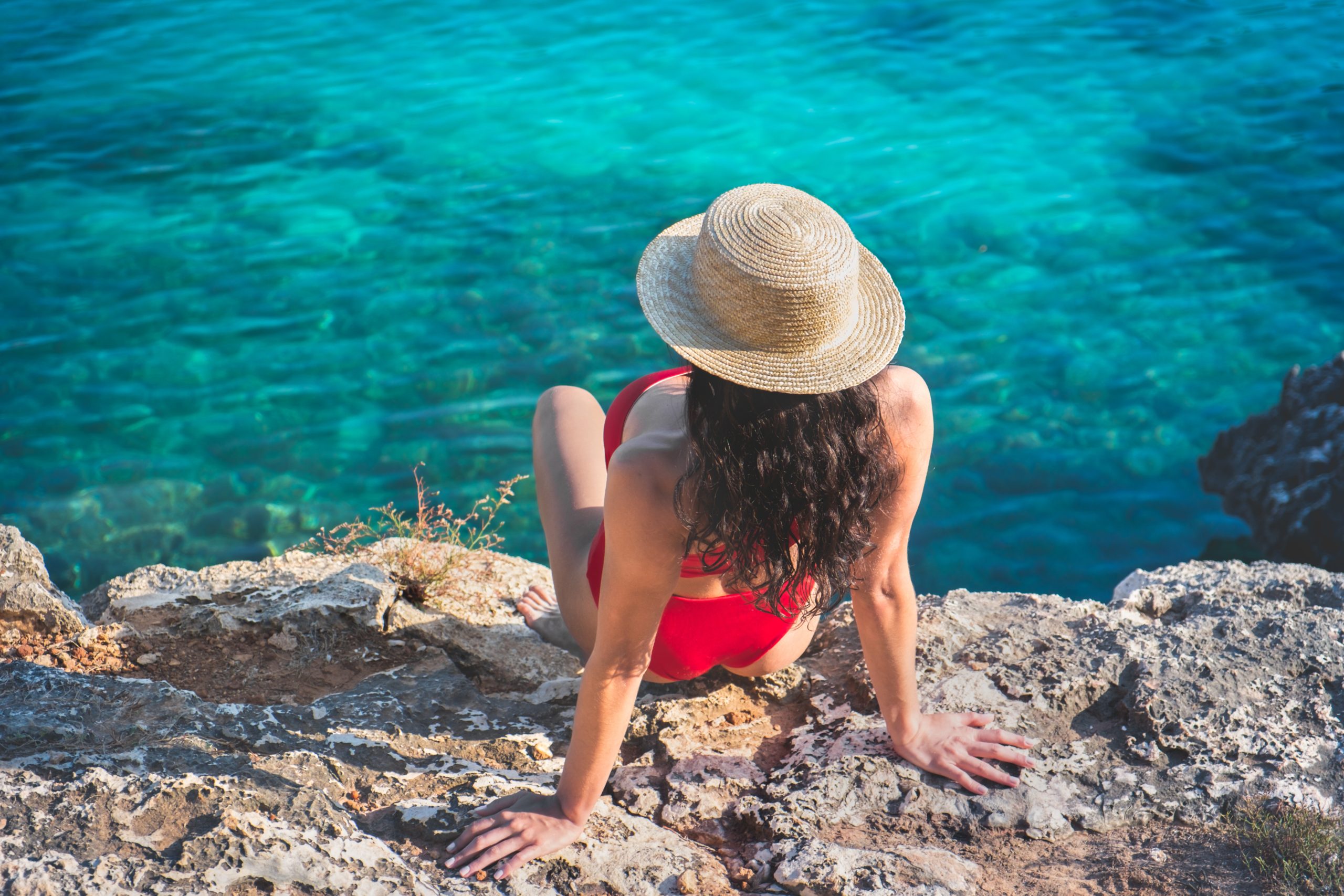 Frau mit Sonnenhut auf einem Felsen am Meer sitzend